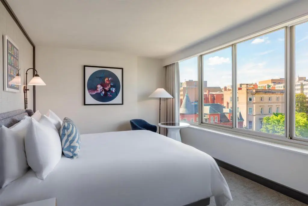 Bright hotel room with a large bed, modern decor, a blue chair, wall art, and large windows overlooking city buildings and trees.