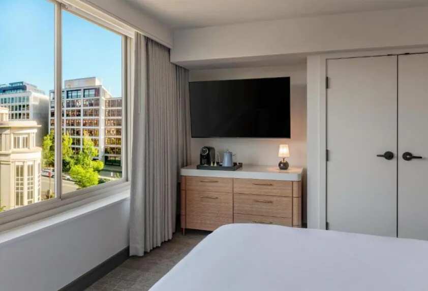 Modern hotel room with a dresser, coffee maker, TV, small lamp, and a large window overlooking city buildings.