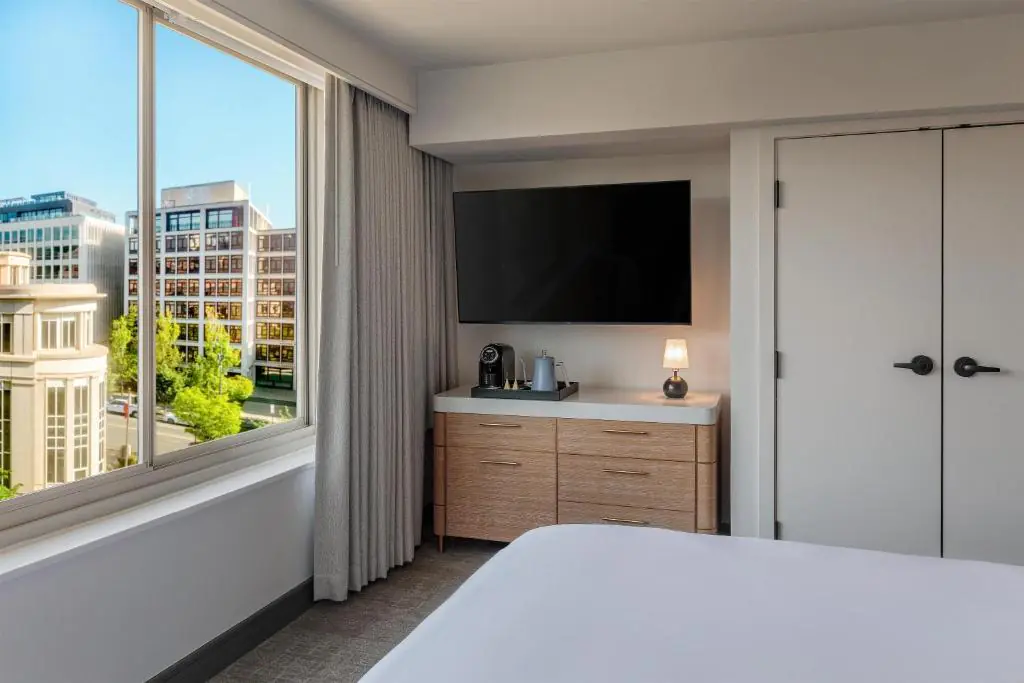 Modern hotel room with a dresser, coffee maker, TV, small lamp, and a large window overlooking city buildings.