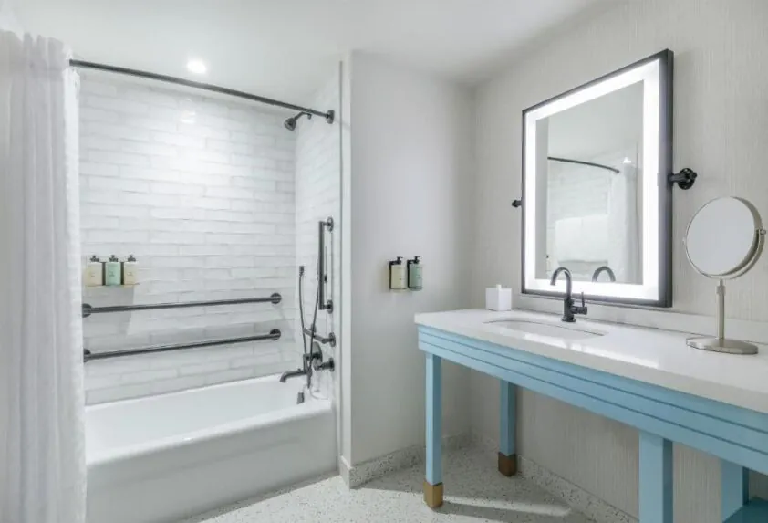Modern bathroom with white tiled shower and bathtub, blue vanity with large mirror, and wall-mounted toiletries. Bright and clean design.