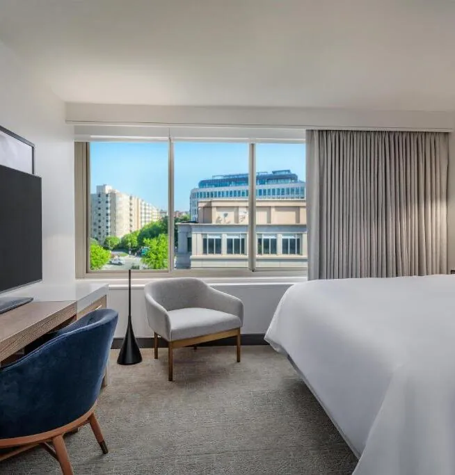 Modern hotel room with a large bed, flat-screen TV, desk, chair, and window with city view and natural light.
