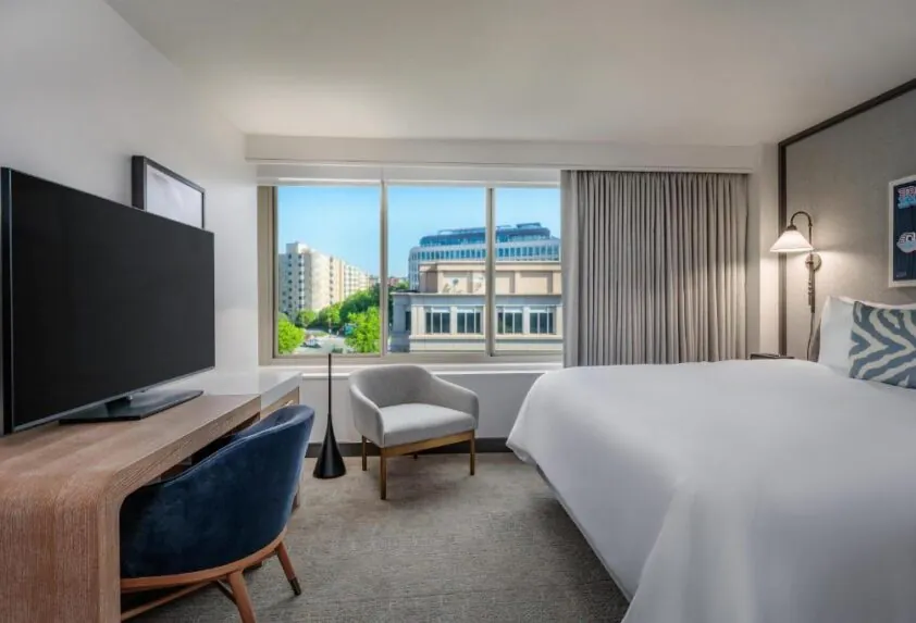 Modern hotel room with a large bed, flat-screen TV, desk, chair, and window with city view and natural light.