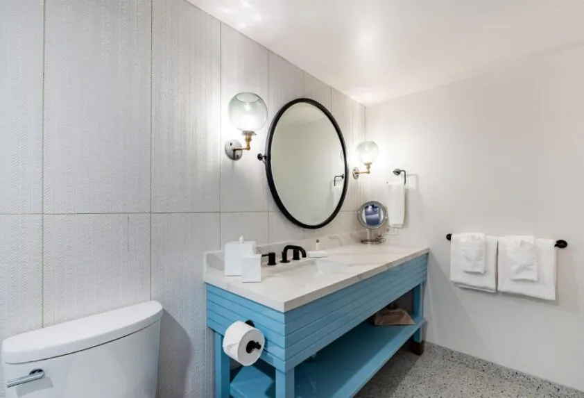 Modern bathroom with a blue vanity, round mirror, wall sconces, white toilet, and neatly folded towels on a rack.