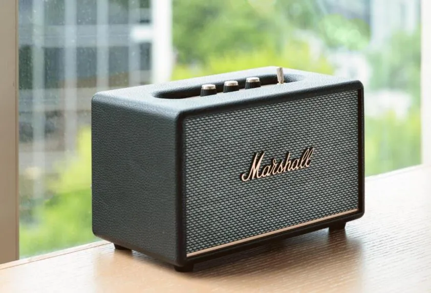 A black Marshall Bluetooth speaker with gold knobs sits on a wooden table by a window with a blurred outdoor view.