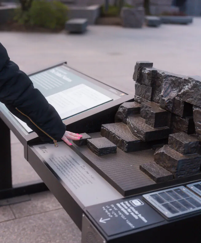 A person touches a tactile display with raised stone steps and braille at an outdoor informational exhibit.