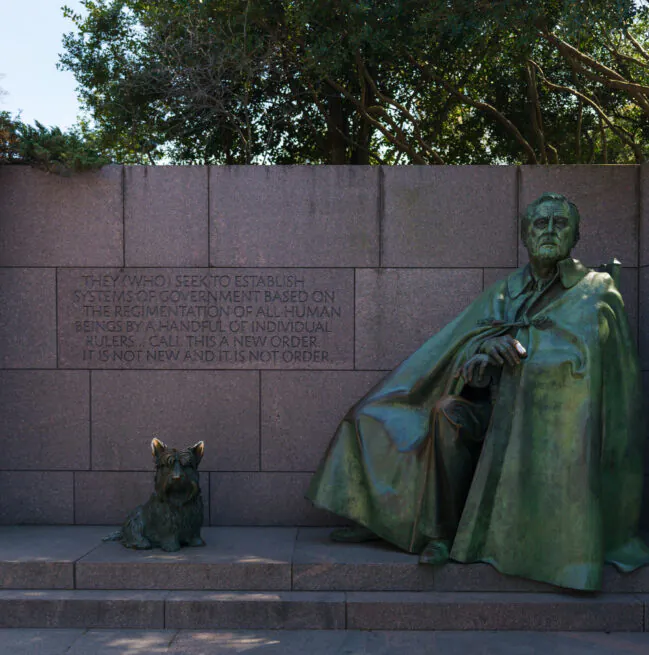 Bronze statue of Franklin D. Roosevelt seated with his dog, Fala, in front of a stone wall engraved with a quote, surrounded by trees.