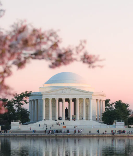 The Jefferson Memorial at sunset with cherry blossoms in the foreground and people gathered near the steps, reflected in the calm water.