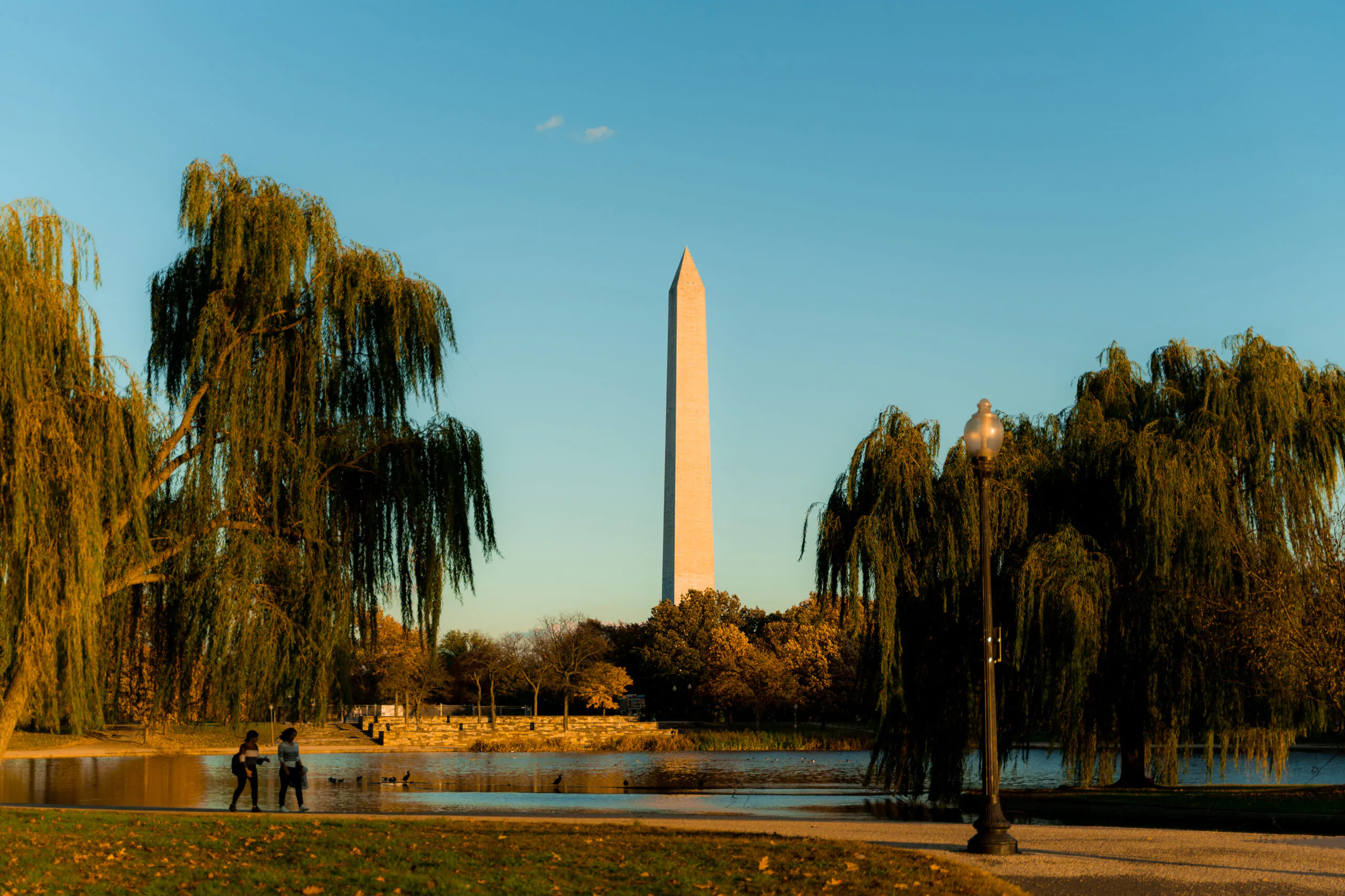 The Washington Monument stands tall at sunset, framed by trees and a pond, with two people walking along the waters edge.
