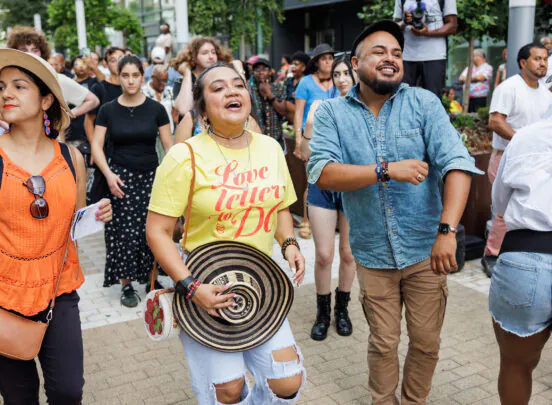 A joyful crowd dances outdoors; a woman in a yellow Love letter to DC shirt smiles, holding a patterned hat, surrounded by people enjoying the event.