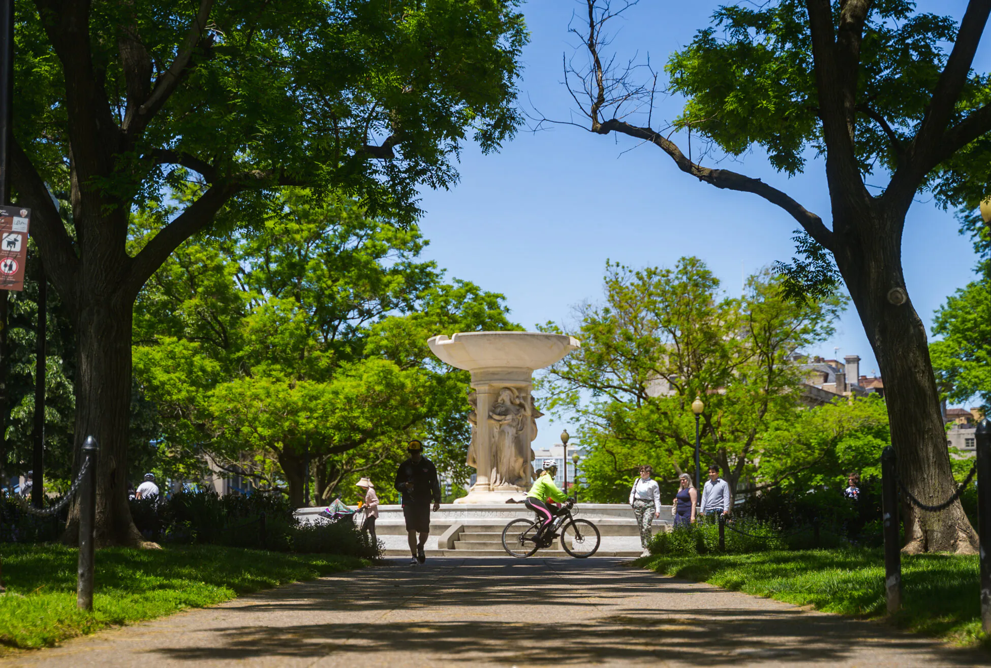 People walk and bike on a tree-lined path toward a large stone fountain in a sunny park, with green foliage and clear blue sky overhead.