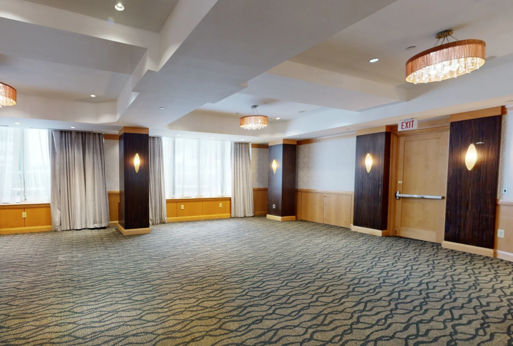 Spacious, empty conference room with carpeted floor, large windows with sheer curtains, wood paneling, and chandeliers.