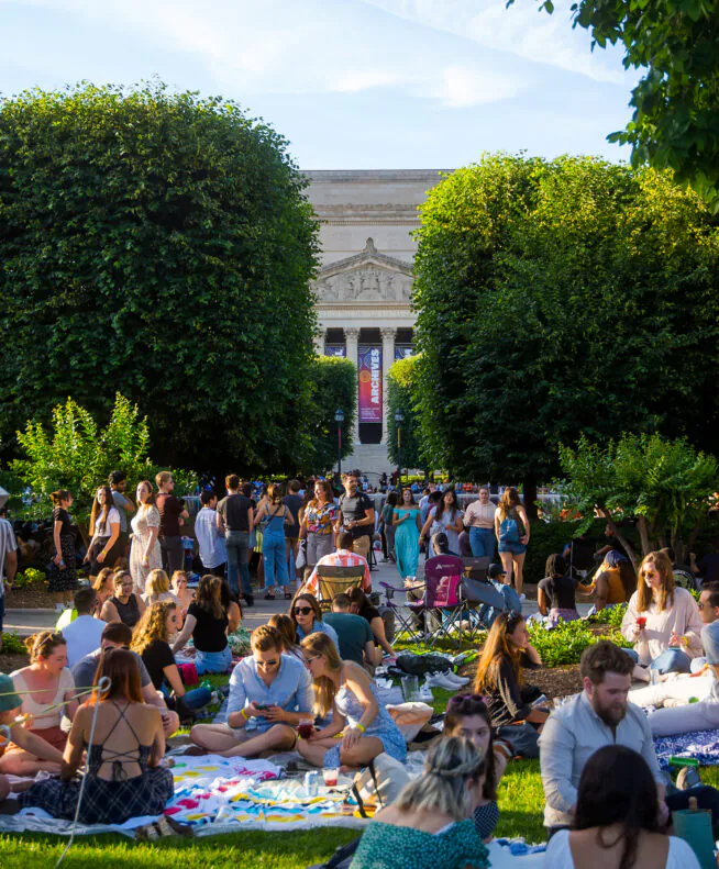 A large group of people having a picnic and socializing on a sunny day in a park with trees and a classical building in the background.
