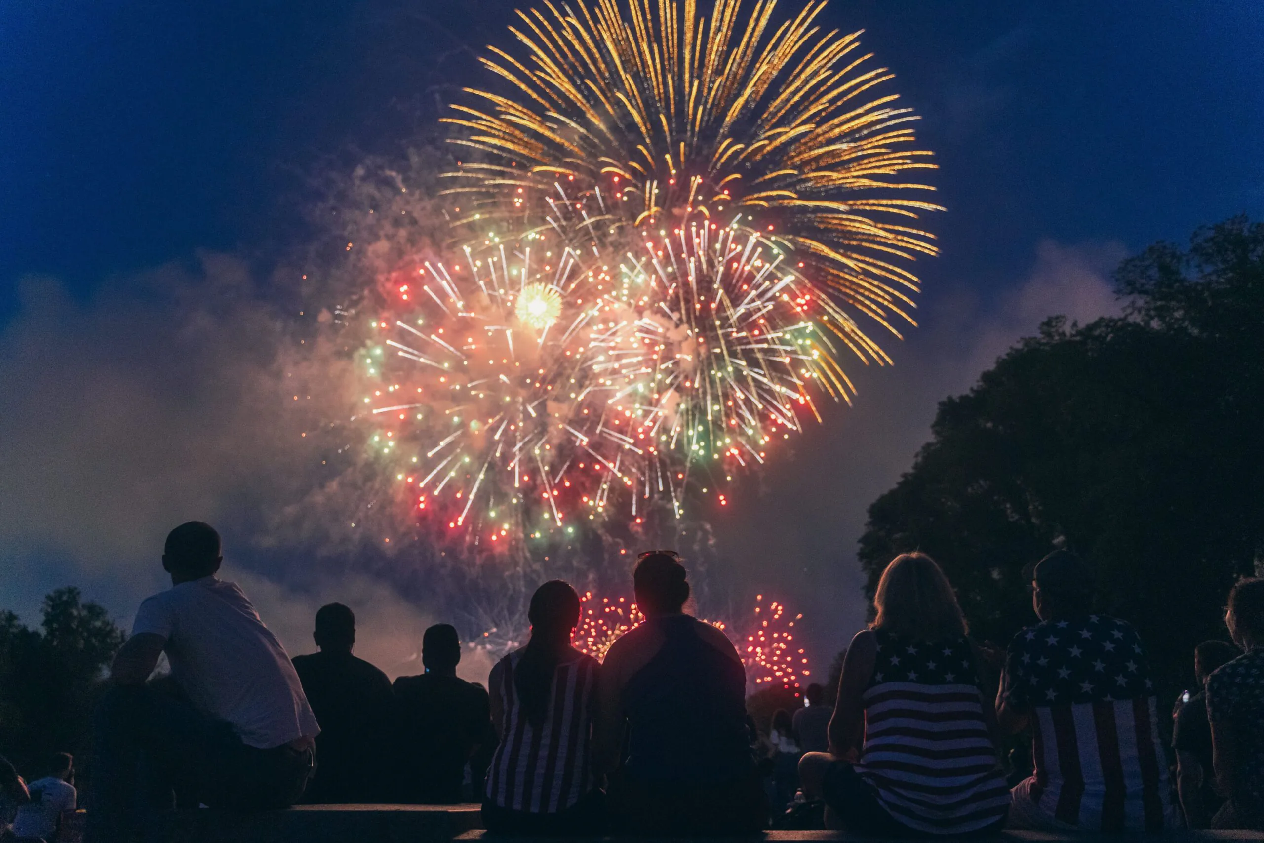 A group of people sit outdoors at night, watching colorful fireworks light up the sky. Some wear clothing with the American flag pattern.