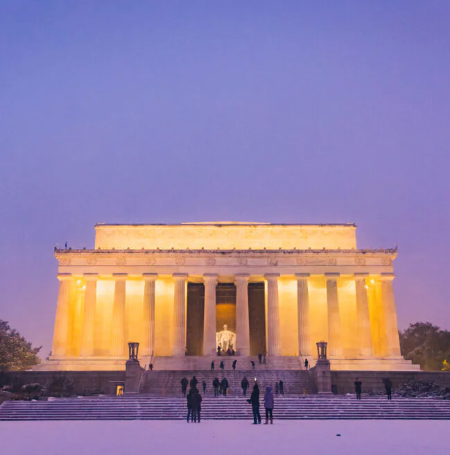 The Lincoln Memorial illuminated at dusk with people gathered on the snow-covered steps and a purple sky overhead.
