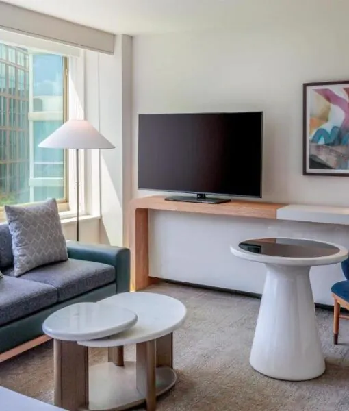 Modern hotel room with large window, sofa, round table, armchair, TV, abstract art, and a view of trees and buildings outside.