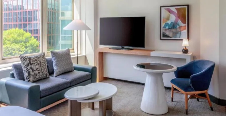 Modern hotel room with large window, sofa, round table, armchair, TV, abstract art, and a view of trees and buildings outside.
