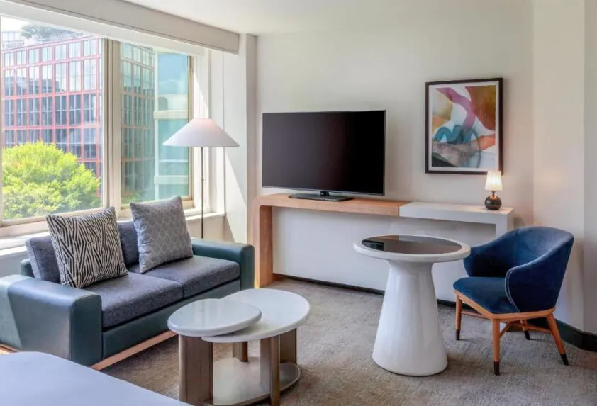 Modern hotel room with large window, sofa, round table, armchair, TV, abstract art, and a view of trees and buildings outside.