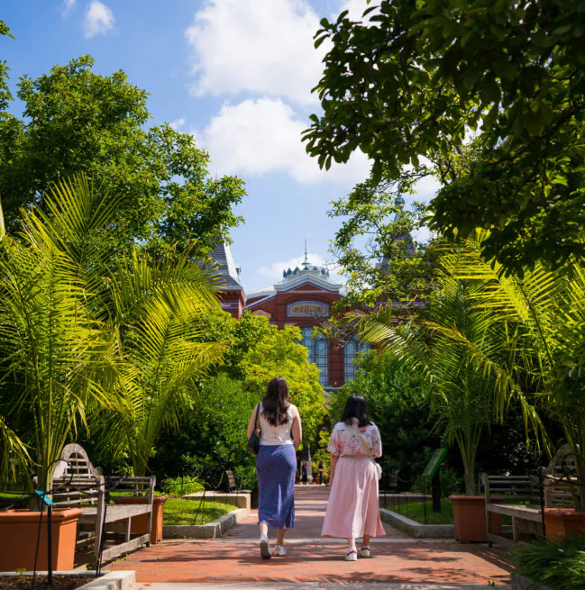 Two women walk along a brick path through a lush garden toward a large, ornate building under a bright blue sky with scattered clouds.