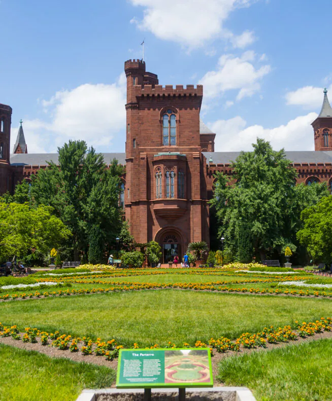 Red brick Smithsonian Castle with towers, surrounded by lush gardens, colorful flowers, and green lawns under a blue sky with scattered clouds.