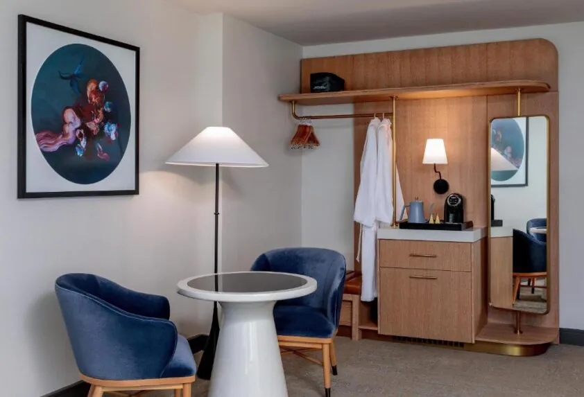 Modern hotel room corner with two blue chairs, a small round table, coat rack, bathrobe, lamp, coffee maker, and colorful artwork on the wall.