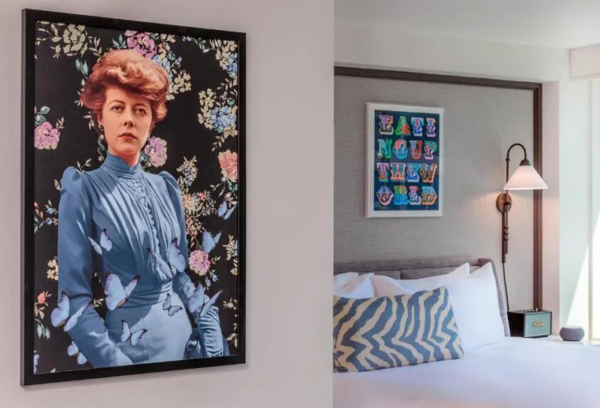 A modern bedroom with a bed, a zigzag pillow, a wall lamp, and two framed artworks, one featuring a woman in blue surrounded by flowers and butterflies.