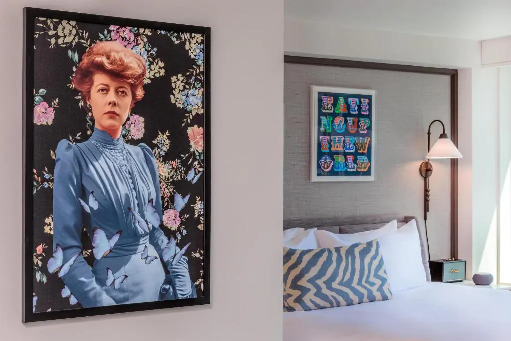 A modern bedroom with a bed, a zigzag pillow, a wall lamp, and two framed artworks, one featuring a woman in blue surrounded by flowers and butterflies.