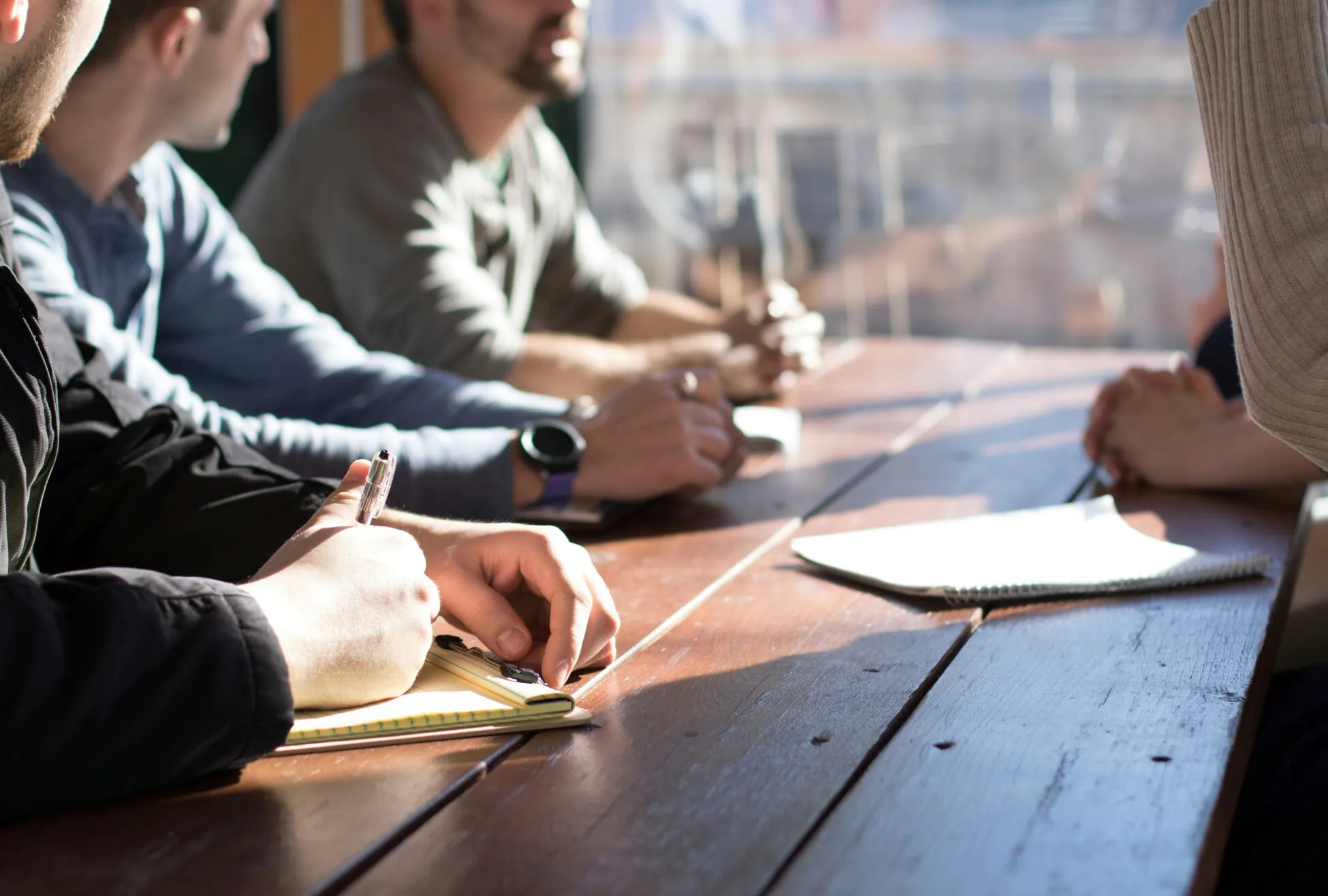 Four people sit at a wooden table in sunlight, holding notebooks and pens, engaged in a discussion or meeting.