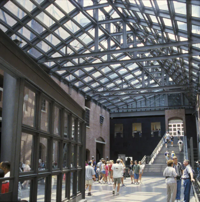 People walk inside a large museum atrium with a glass roof and brick walls; a staircase is visible in the background.