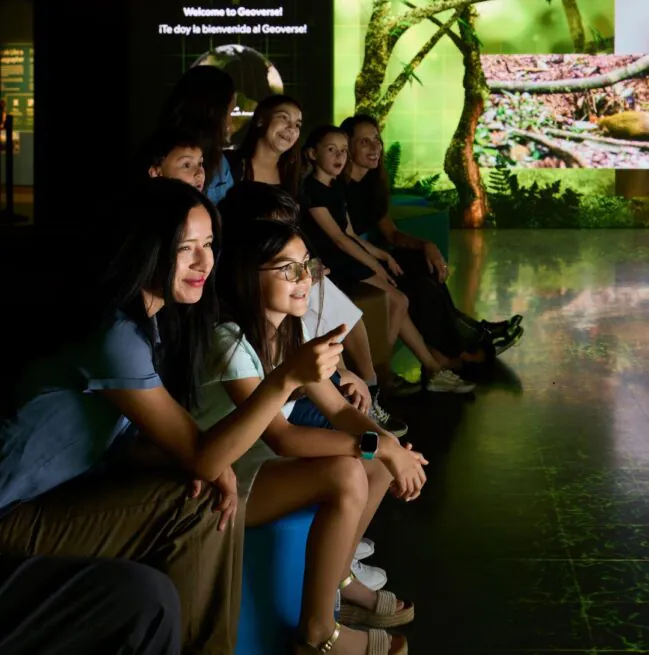 A group of people sit on benches, smiling and watching an exhibit at a National Geographic display with nature images in the background.