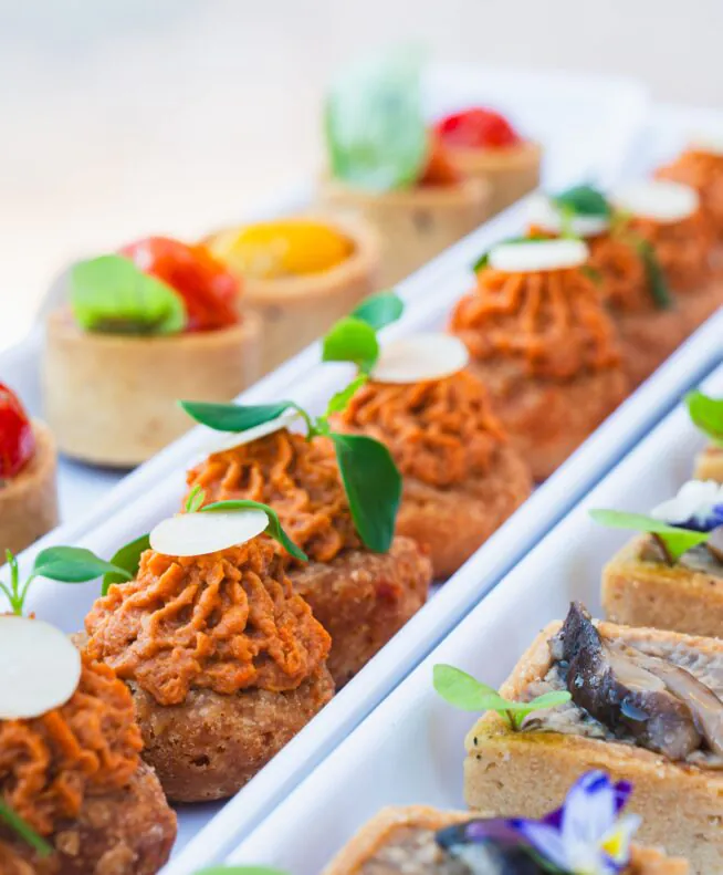 Rows of assorted bite-sized appetizers garnished with greens and edible flowers, arranged neatly on a white rectangular platter.