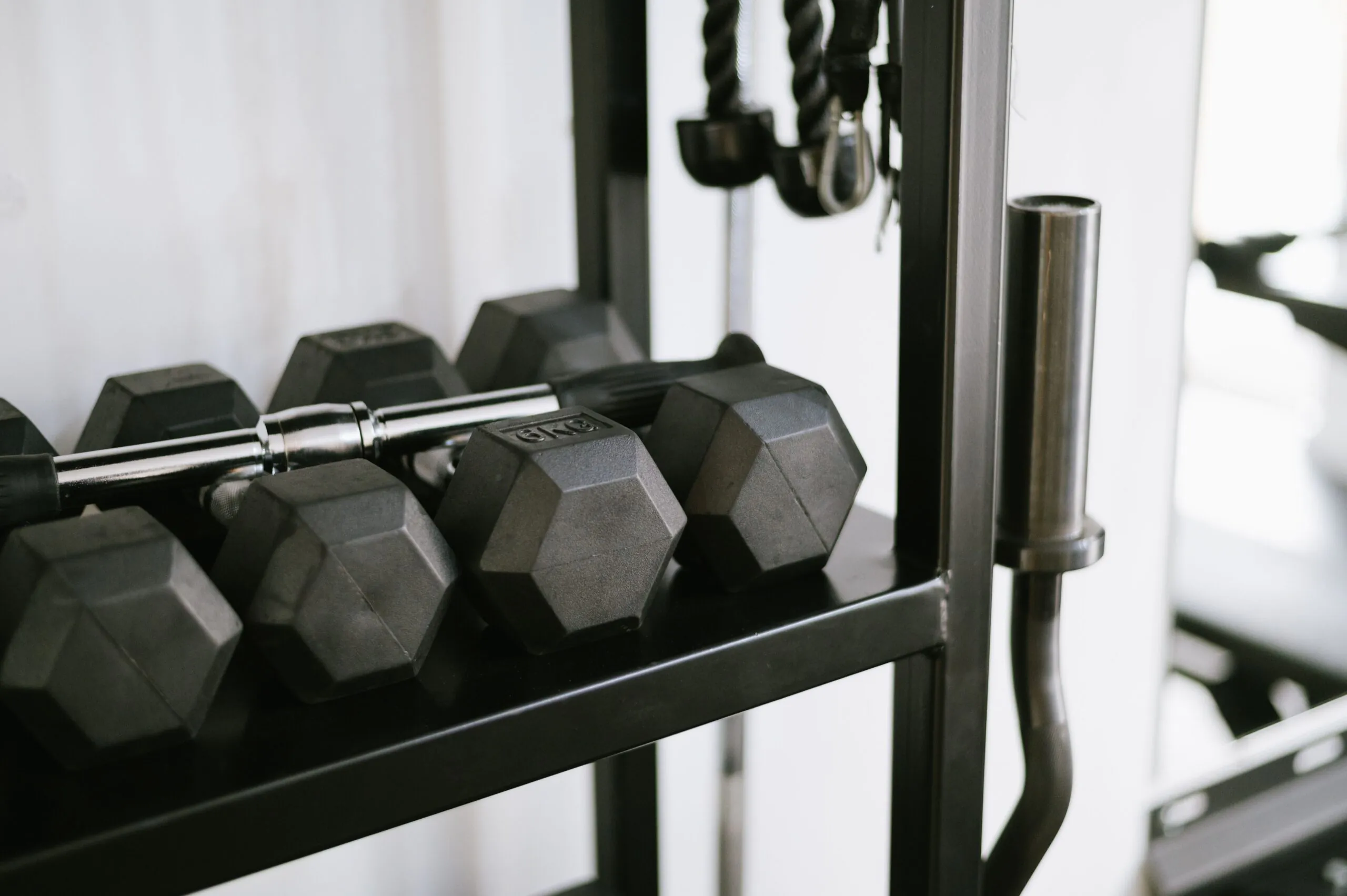 Black hexagonal dumbbells and a barbell on a rack in a gym with workout equipment visible in the background.