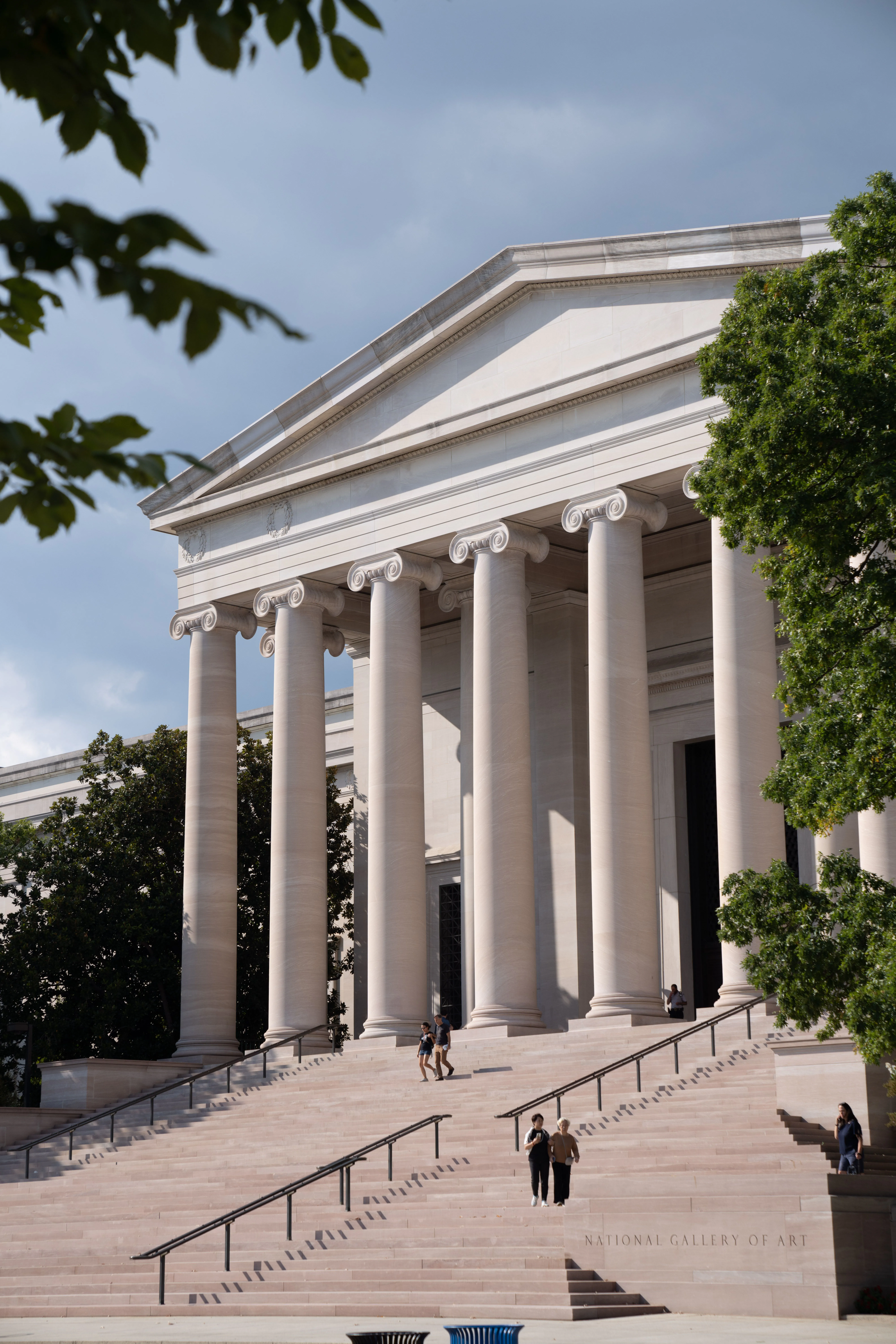 People walking up steps toward a large neoclassical building with tall columns, surrounded by green trees; text reads National Gallery of Art.