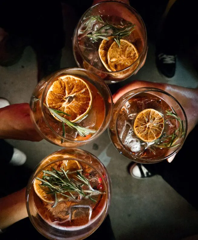 Four people holding glasses with cocktails garnished with dried orange slices and rosemary, viewed from above against a dark background.