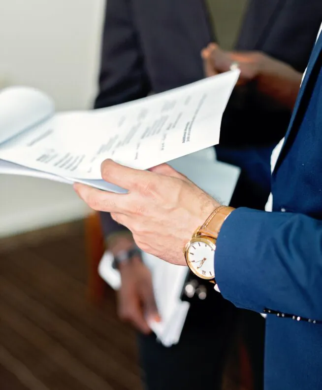 Close-up of a person in a blue suit holding documents, with another person in the background also holding papers.
