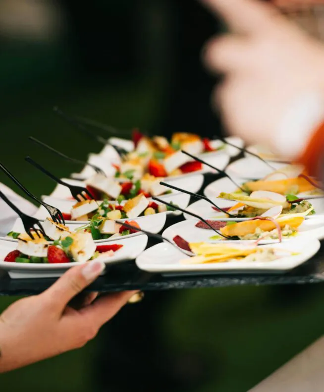 Person holding a tray of assorted small appetizers with forks, while others reach for the food at what appears to be an event or gathering.