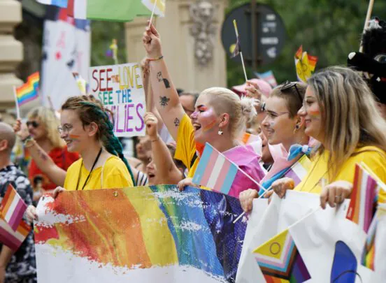 A diverse group of people at a pride parade hold LGBTQ+ flags and banners, smiling and celebrating together.