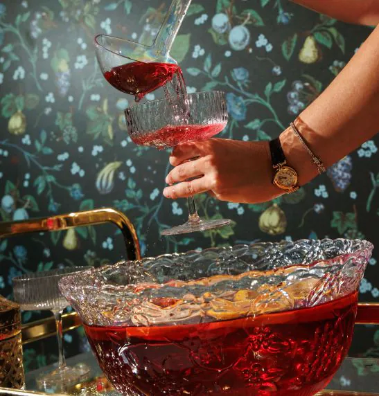 A person pours red punch from a glass cup into another, above a crystal punch bowl with fruit, in front of a floral-patterned wall.