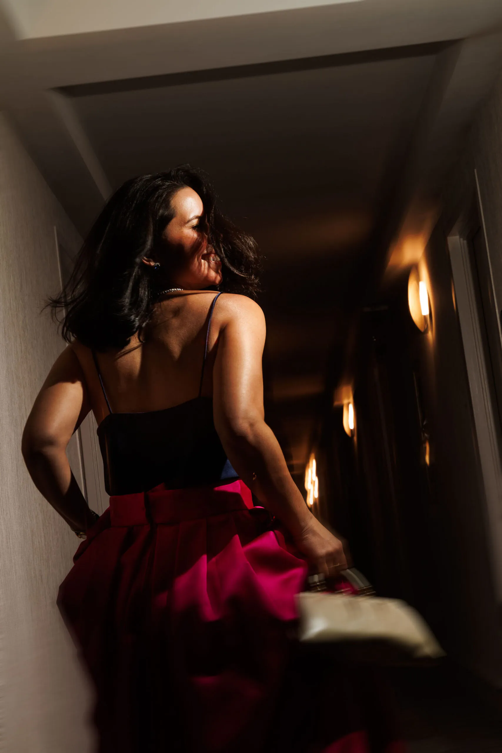 A woman in a black top and pink skirt stands in a dimly lit hallway, looking over her shoulder and holding a small handbag.