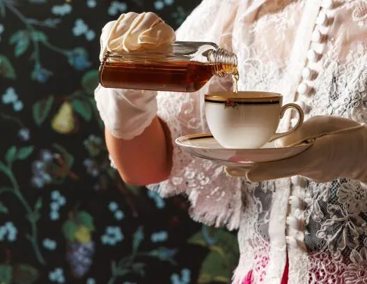 A person in lace gloves pours liquid from a glass bottle into a teacup, with a floral patterned background.
