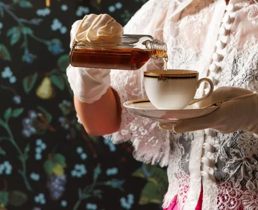 A person in lace gloves pours liquid from a glass bottle into a teacup, with a floral patterned background.