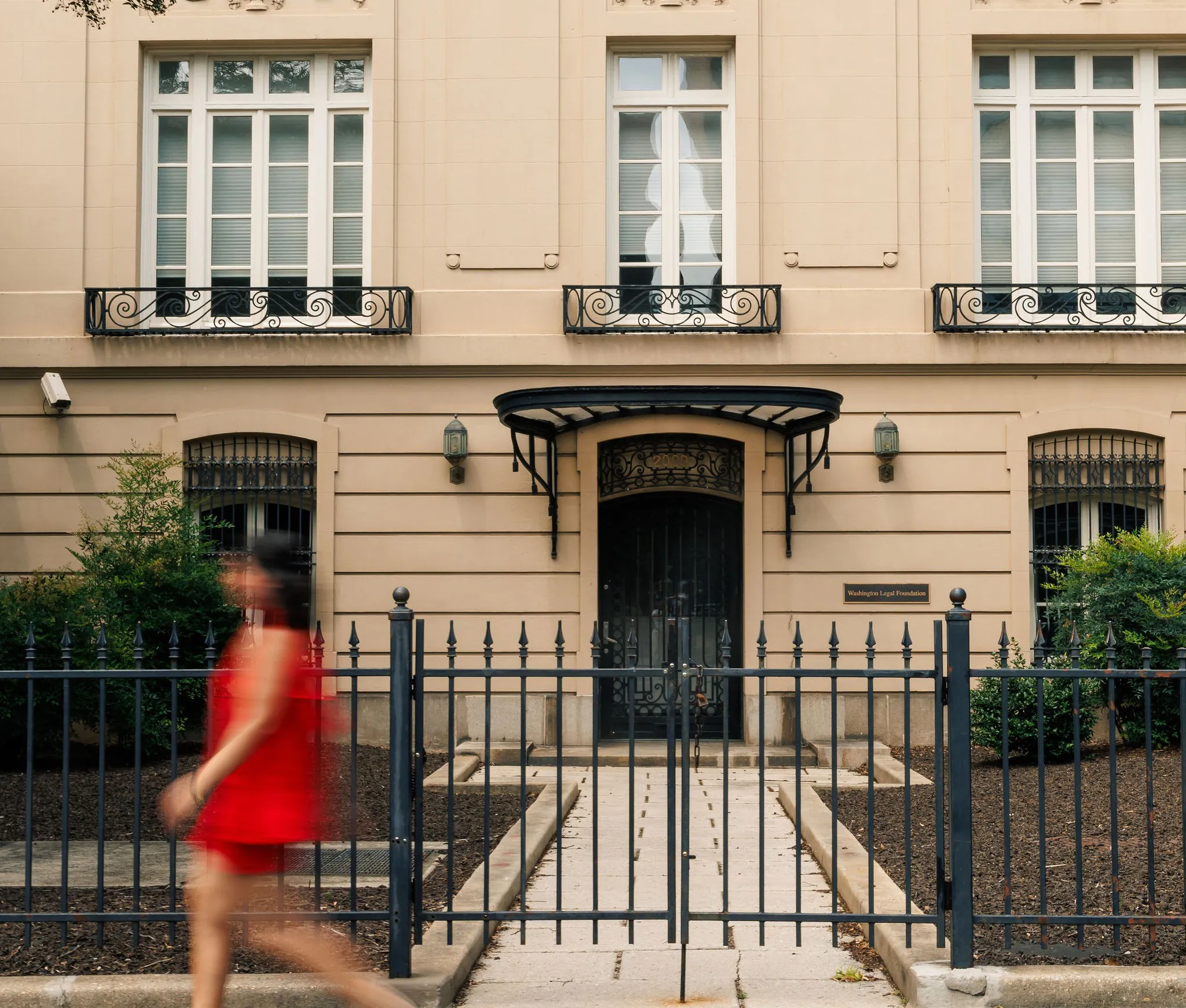 A person in a red outfit walks past a beige building with tall windows, black railings, and a gated entrance.
