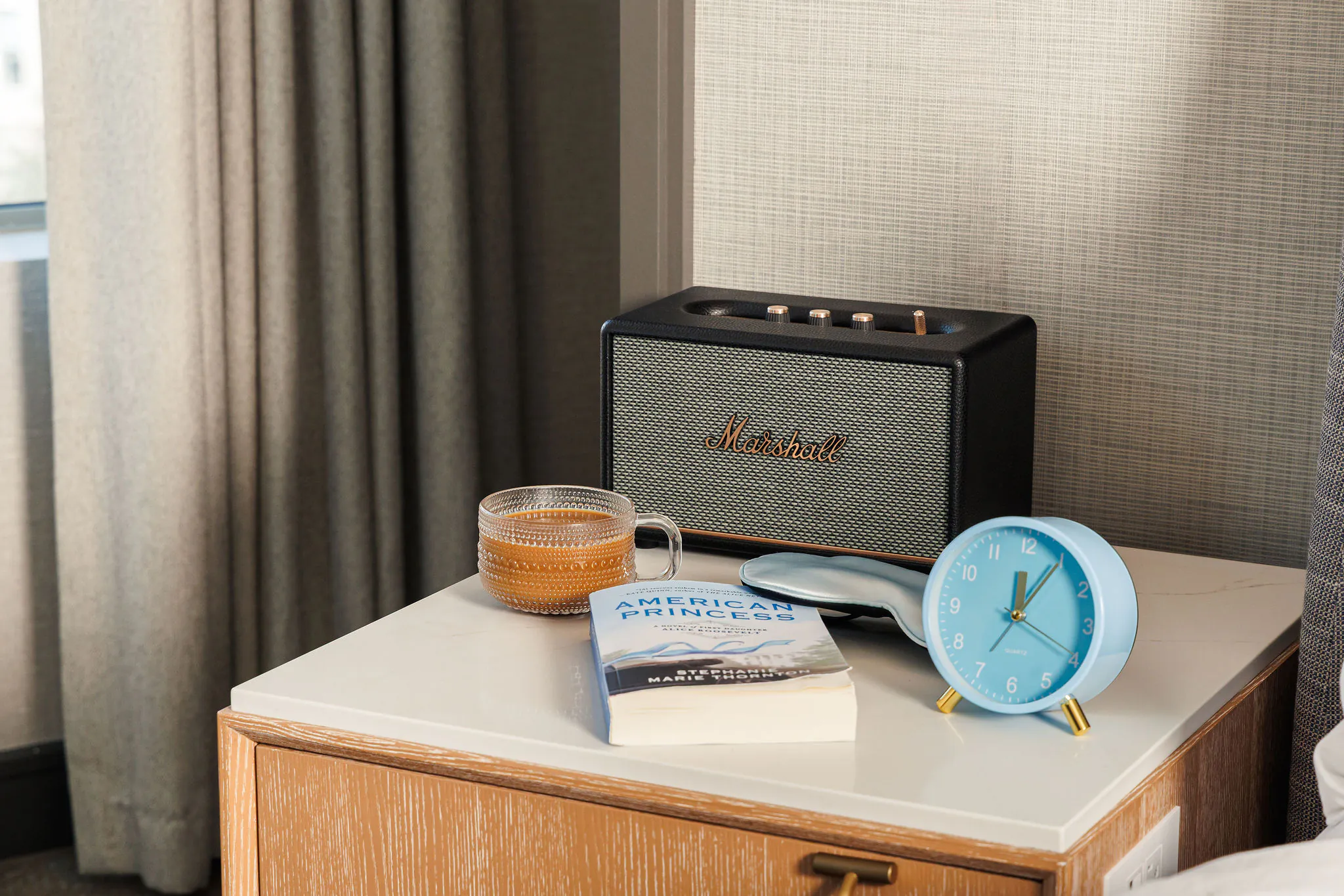 A book, teacup, blue alarm clock, sleep mask, and Marshall speaker sit on a bedside table next to a window with curtains.