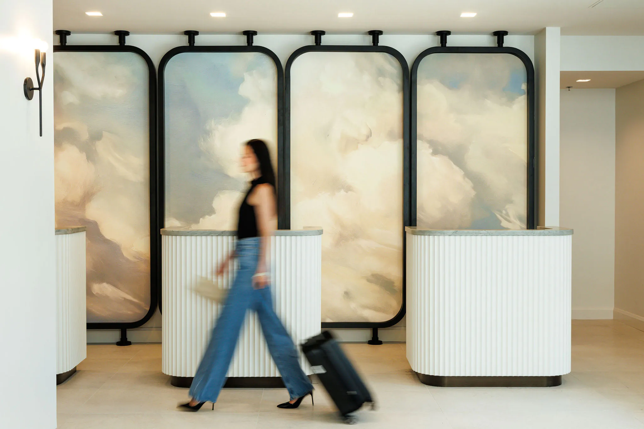 A woman in jeans walks with a suitcase past modern, white reception desks and a cloud-themed wall mural in a bright lobby.