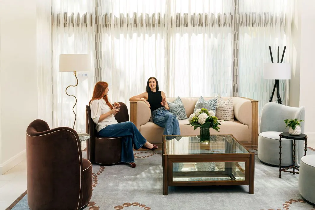 Two women sit and chat in a bright, modern living room with neutral furniture, large windows, and a glass coffee table with flowers.