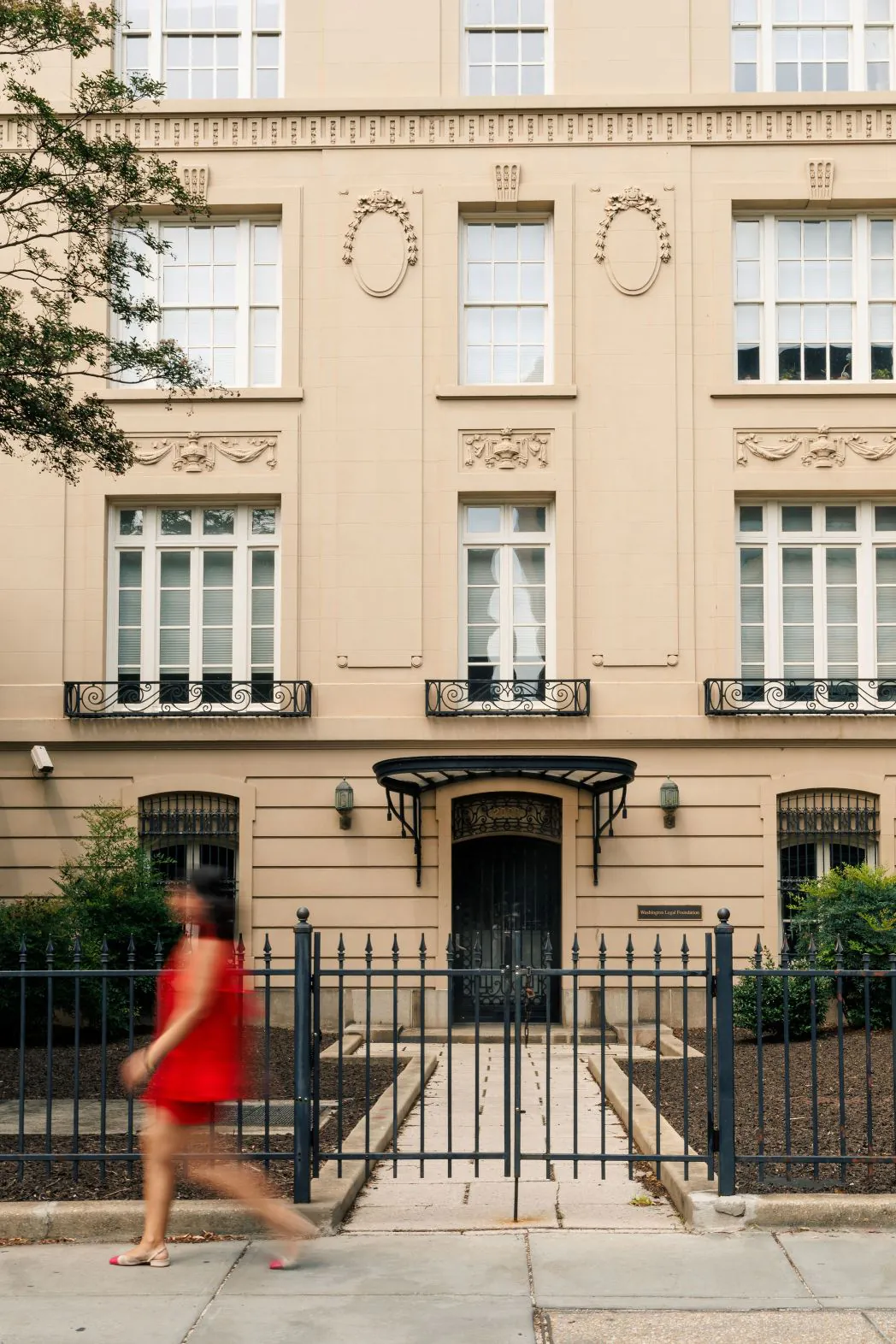 A woman in a red dress walks past a beige, ornate building with large windows and black iron fencing.